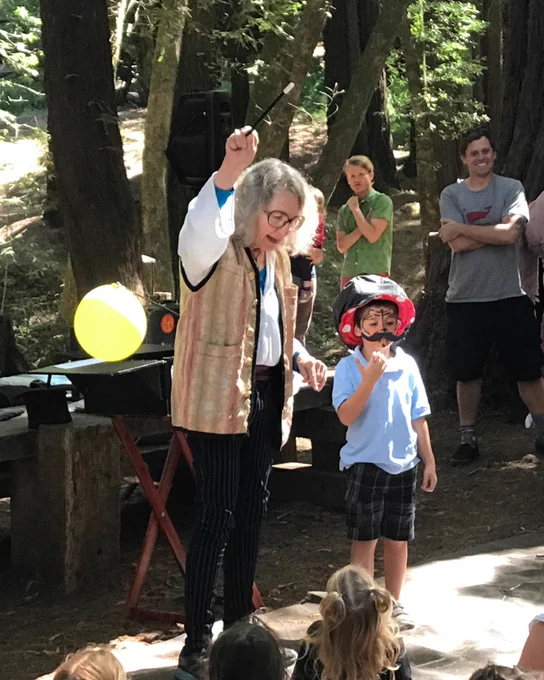 Outdoors, surrounded by people and redwood trees, a magician stands with wand raised as a young volunteer in a helmet stands next to her.