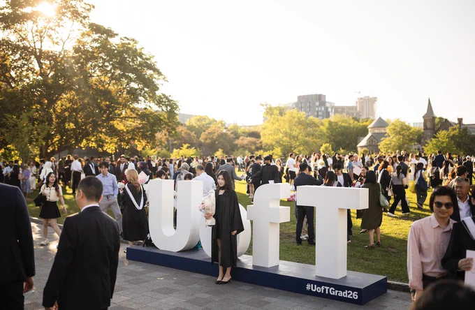 A person in academic regalia stands next to large "U of T" letters at an outdoor graduation ceremony. Many people are gathered in the background on a sunny day. Trees and buildings are visible.
