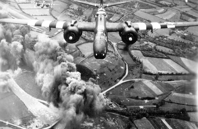 A black and white photograph of a World War II bomber, likely a B-25 Mitchell, flying low over a rural landscape with visible smoke plumes rising from the ground.