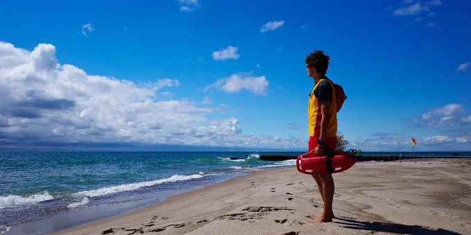 Lifeguard in orange vest holding rescue buoy, standing barefoot on sandy beach looking at the ocean under a partly cloudy sky.