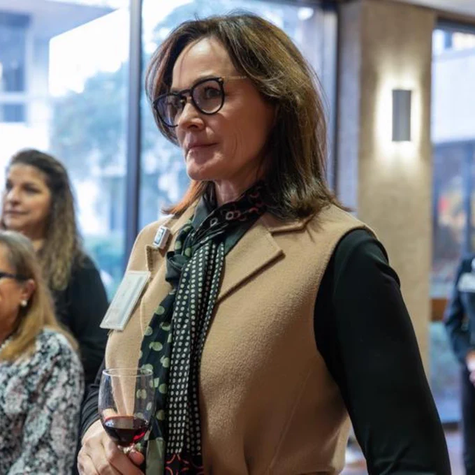 Woman wearing a tan vest and patterned scarf holding a glass of red wine at an indoor social event.