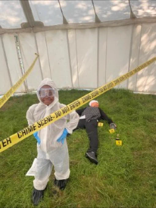 Child in white overalls dressed as a forensic investigators in an obviously fake crime scene.