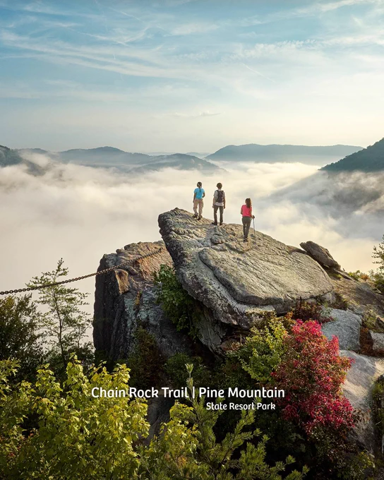 Three individuals stand on a large rock outcropping on the Chain Rock Trail at Pine Mountain State Resort Park. They overlook a foggy valley surrounded by distant mountains. Lush green foliage and colorful shrubs are visible in the foreground.