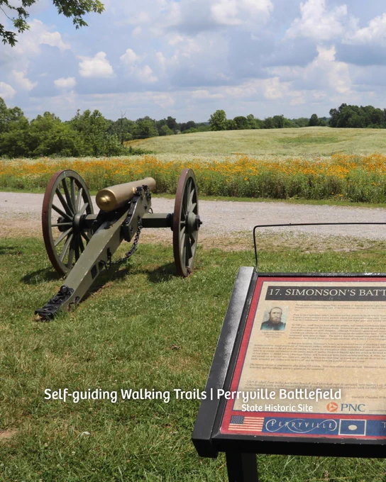 A historic cannon sits on a grassy area near a path surrounded by wildflowers at Perryville Battlefield. A nearby informational plaque is titled "Simonson's Battery," providing historical details about the site.