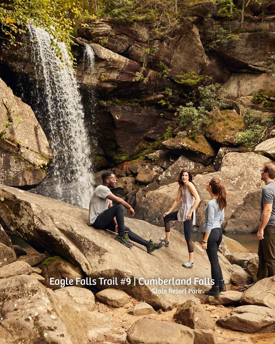 A group of people rests on large rocks near a waterfall at Eagle Falls Trail #9, Cumberland Falls, in Stoke Resort Park. The waterfall cascades down a rocky cliff surrounded by greenery.