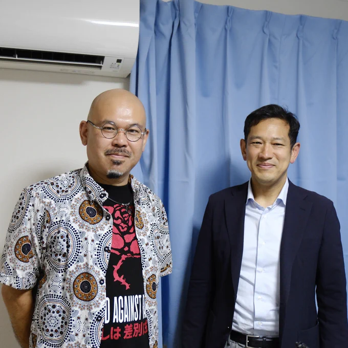 左側がゲストの植田さん。
丸眼鏡をかけ、幾何学模様の入った開襟シャツの下には、赤い薔薇のイラストと文字が描かれた黒いTシャツを着用している。

右側が宮本徹元衆議院議員。
紺色のジャケットと淡い色のシャツを着て穏やかな笑顔をしている。