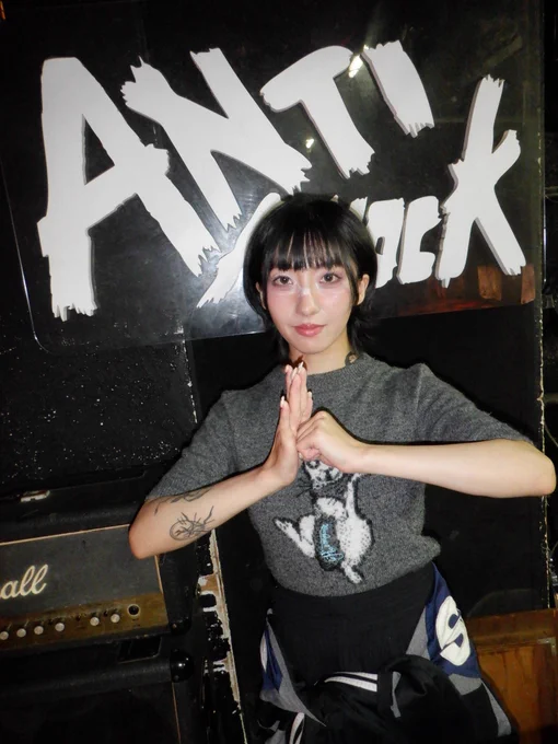 A person in a grey knit top with an animal graphic and a patterned skirt presses their palms together in a prayer-like pose in front of the ANTIKNOCK live venue sign. A Marshall amplifier is visible to the left, and white face paint is on the nose bridge.

アニマルグラフィックのグレーのニットトップスと柄スカートを着た人物が、ANTIKNOCKのライブハウスの看板の前で両手を合わせたポーズをとっている。左にはMarshallのアンプ、鼻の付け根には白いフェイスペイント。