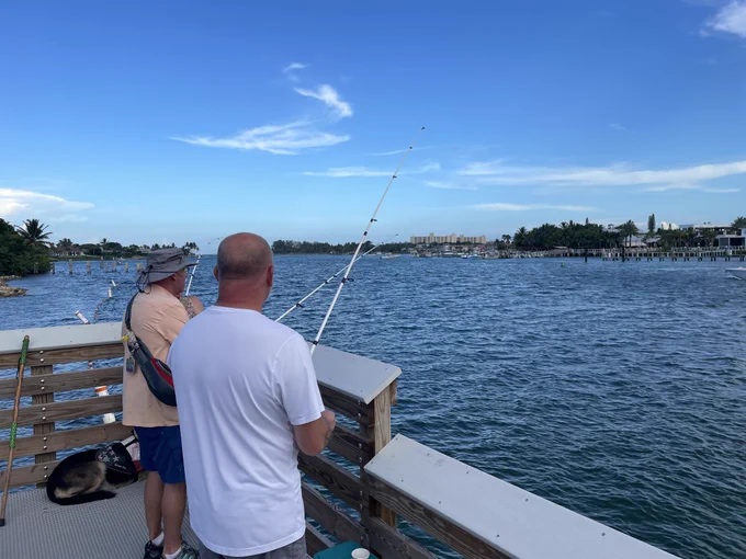 Two men fishing off a wooden pier overlooking a calm blue bay under a clear sky.