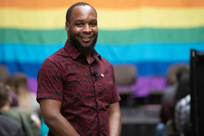 A man stands in front of a multicolored flag