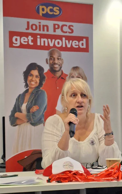 The photo shows PCS DWP President Angela Grant holding a microphone with a backdrop of a PCS pull-up stand with the words join PCS, get involved.