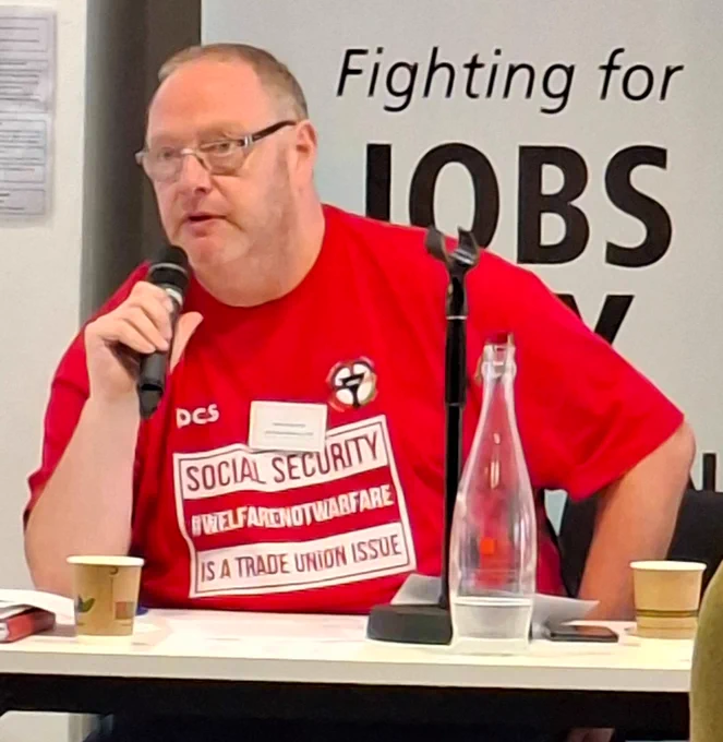 The photo shows PCS DWP Group Deputy President Martin Cavanagh holding a microphone and wearing a T-shirt with the words social security is a trade union issue, #WelfareNotWarfare and with a partially obscured backdrop which reads fighting for jobs