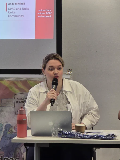 The photo shows Ellen Morrison, DPAC Executive Committee and Unite member sat at a desk holding a microphone with a laptop in front of her.