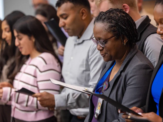 A group of people stands closely together, each holding papers. They appear focused and engaged in an activity, possibly reading or singing. The individuals are dressed in formal or office attire, and the setting has a professional atmosphere. One person in the foreground wears glasses and has braided hair, concentrating on the paper they hold.
