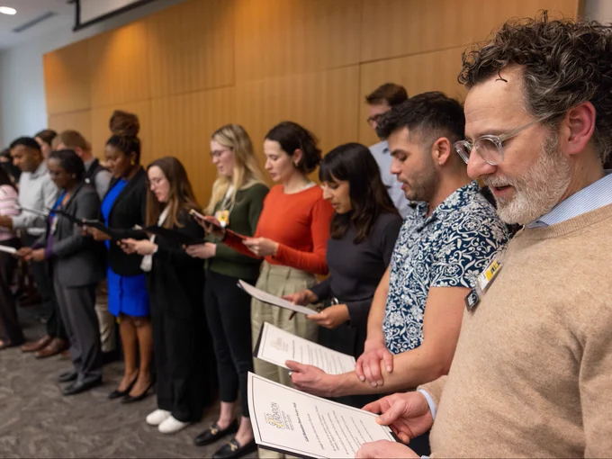 A diverse group of people stands in a conference room, reading from printed documents. They are lined up side by side, focusing intently on the papers in their hands. The setting is a professional meeting or ceremony, with a wooden-paneled wall in the background.