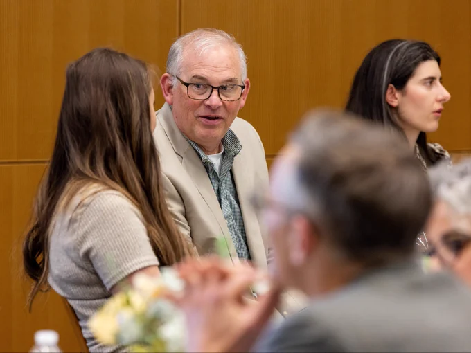A group of people is seated in a room with wooden paneling. A man wearing glasses and a beige blazer is engaged in conversation with a woman with long brown hair. Another woman with long dark hair is visible in the background, looking away. The foreground is blurred with a focus on the people in the middle.