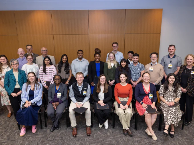 A diverse group of people is posed in a well-lit room with a wooden panel wall background. They are arranged in two rows, with some seated in the front and others standing behind. Many are smiling, dressed in a mix of business casual attire, and some hold black folders and wear ID badges. The atmosphere appears professional and friendly.