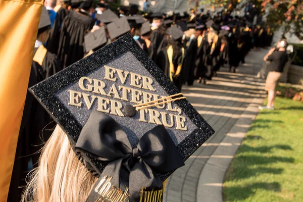 A person wearing a graduation cap decorated with the words "Ever Grateful, Ever True" is in the foreground. In the background, many graduates in caps and gowns are walking along a curved path.