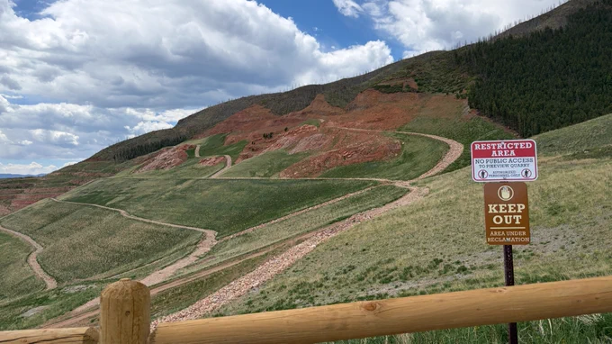 A landscape of rolling hills with a mix of green grass and red soil. Paths wind through the terrain. A wooden fence in the foreground has signs reading "Restricted Area" and "Keep Out Area Under Reclamation." A forested area and a cloudy sky are visible in the background.