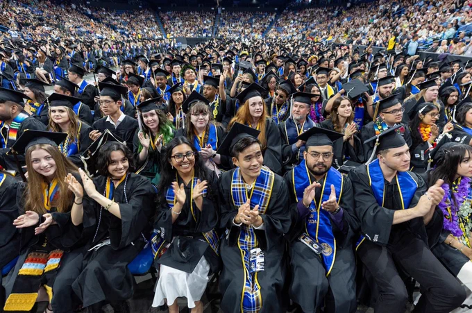 Graduates sitting in an arena in caps & gowns clapping