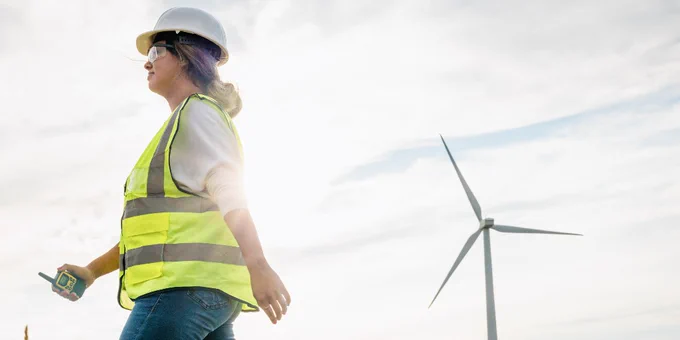 A person in a high-visibility vest and helmet is walking while holding a radio. There is a wind turbine in the background against a cloudy sky.