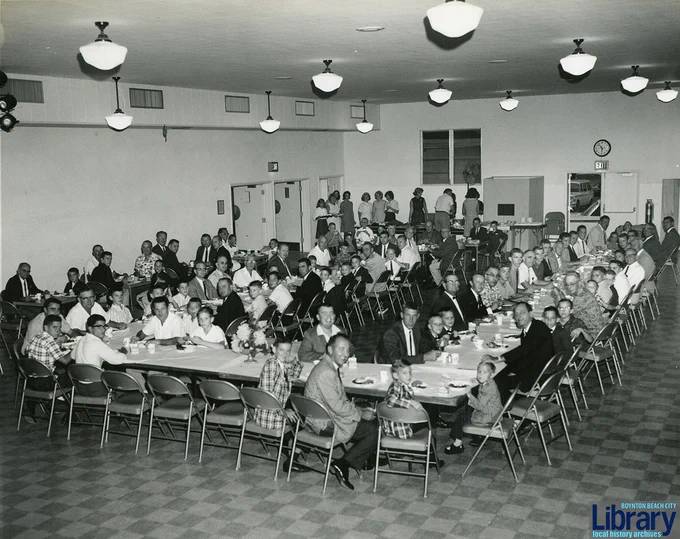 A large group of men, women, and children seated around long tables in a spacious, well-lit hall, engaged in a communal meal or gathering, with some people standing and conversing near the back of the room.