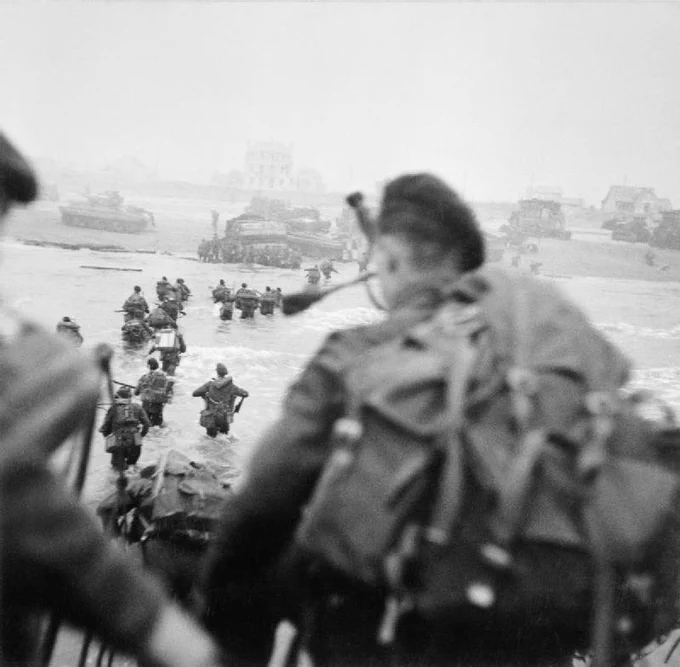 This is a historical black and white photo depicting the D-Day landings during World War II. The image shows soldiers disembarking from landing crafts and moving toward the beach. In the foreground, a soldier is seen from the back, carrying military gear. The background features multiple soldiers wading through water, with buildings visible along the shoreline under overcast skies. No text is visible in the image.