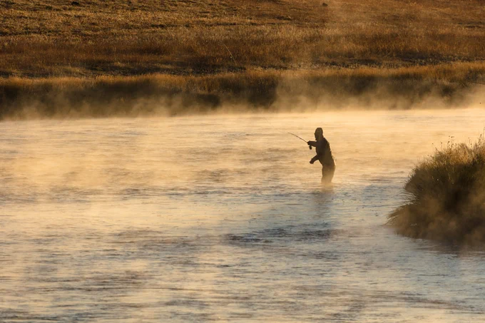 A person standing knee-deep in a river at dawn. The person is casting a fishing line into the water. Steam can be seen rising from the river.  