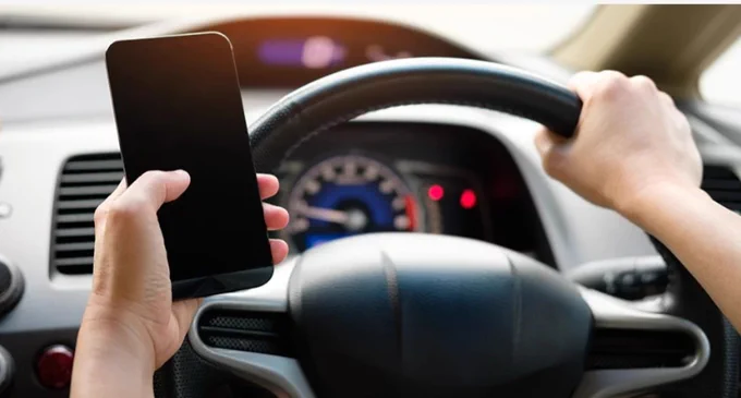 A person is driving, holding a smartphone in one hand and steering the car with the other. The vehicle's dashboard and speedometer are visible.