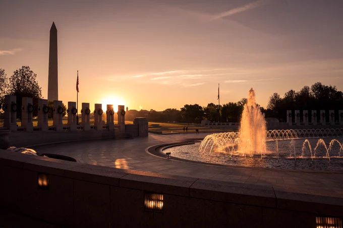 The World War II Memorial in Washington, D.C., at sunrise. The sun rises just beyond the pillars and flagpole, casting a golden glow across the plaza. A large fountain is in the foreground and the Washington Monument stands tall in the background. 