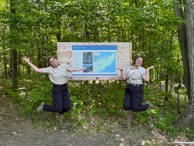 Two Ontario Parks staff members jumping in front of trailhead sign. 