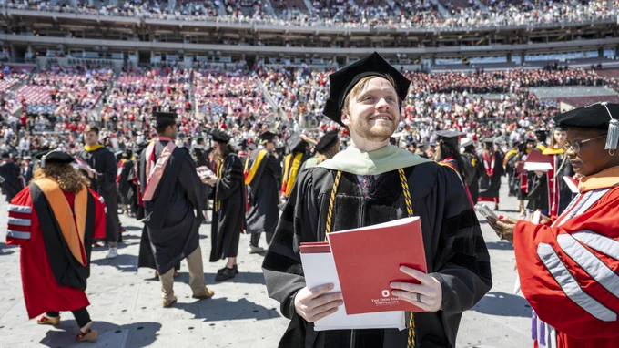 A graduate in academic regalia smiles while holding a red diploma cover at a ceremony in a stadium filled with seated attendees. Other graduates and officials are present, wearing gowns and hoods in various colors associated with The Ohio State University.