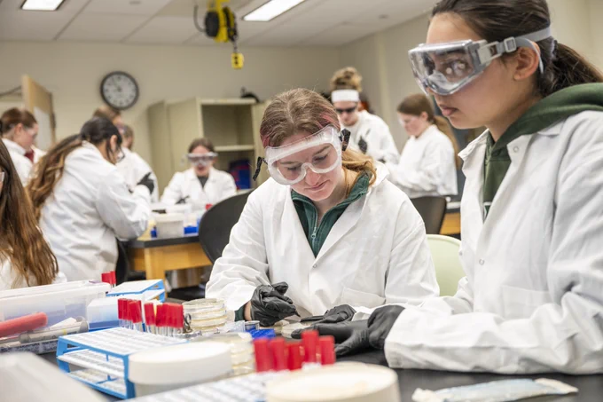 A group of students in a laboratory wearing protective goggles and white lab coats are working at a table with various lab equipment, including petri dishes and test tubes.