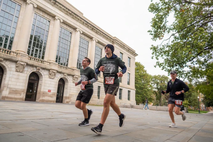 Three people jogging on a paved path in front of Thompson Library. Each wears a race bib. Trees and greenery are visible in the background, with a bright, cloudy sky above.