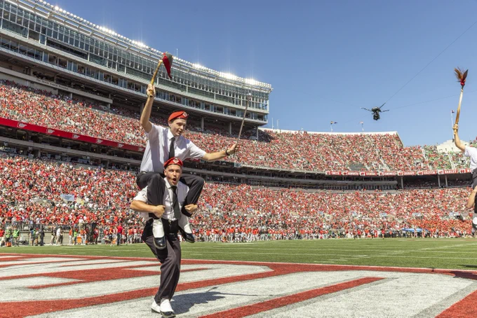 A crowded football stadium filled with fans wearing red. Two individuals in white shirts and suspenders, each holding red objects, are standing on the field. One is sitting on the shoulders of the other. The audience is in the background, with bright stadium lights above.