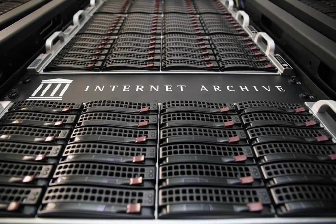 Close-up photo of Internet Archive server racks, showing rows of hard drives with red pull tabs and the Internet Archive logo and name on a black panel.