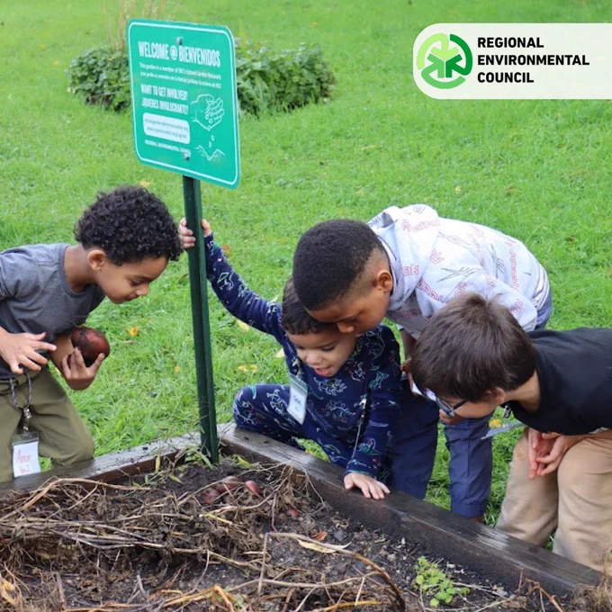Four children closely examine a raised garden bed with soil and plants, next to a bilingual welcome sign.