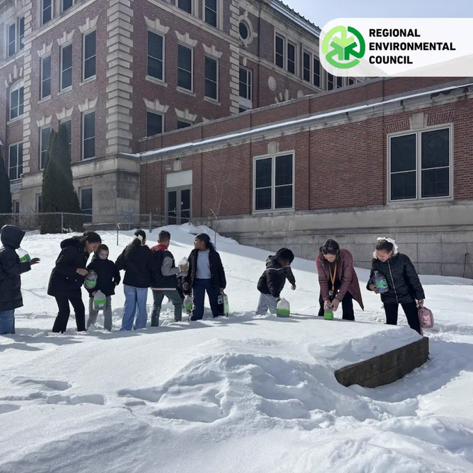 Group of children putting out home-made greenhouses at a brick school building on a sunny winter day.