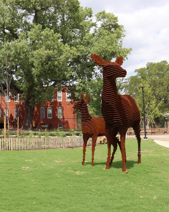 Two rust-colored metal deer sculptures stand on a grassy lawn near a large tree and red brick building under a partly cloudy sky.