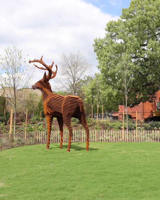 Rust-colored metal sculpture of a stag with large antlers standing on a green lawn near a wooden fence and trees.