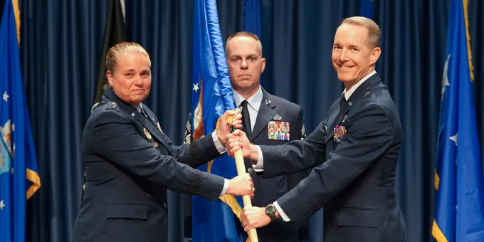 Three uniformed military personnel hold a ceremonial flag together during a formal event with blue curtains and flags in the background.