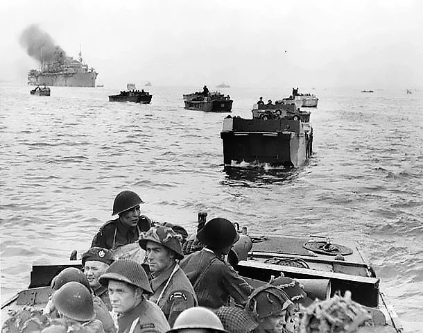 Black and white historical photo of Allied soldiers in landing craft approaching a beach during World War II, with ships visible in the background.