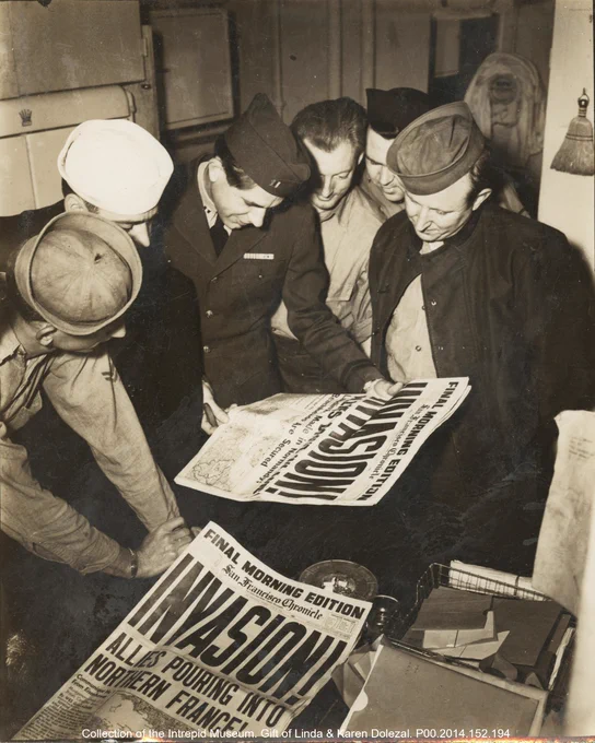 Four individuals in military attire closely examine a newspaper headlined "Invasion Starts," indicating a historical wartime event. The setting appears to be indoors, possibly a military facility.