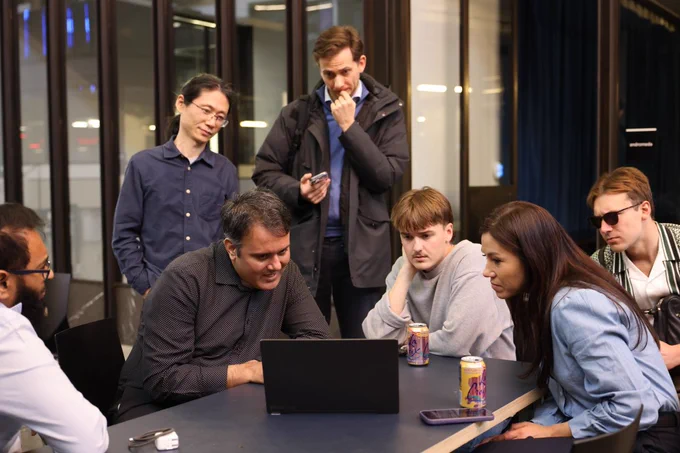 A group of people gathered around a table, looking at a laptop. One person in a dark shirt is seated and focused on the screen. Others stand or sit close by, intently observing, while some hold drinks. The setting appears to be a modern, indoor environment with large windows.