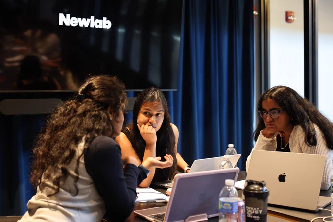 A group of three people sit at a table with laptops open, engaged in discussion. In the background, a screen displays the logo of "Newlab." The setting suggests a collaborative work environment.