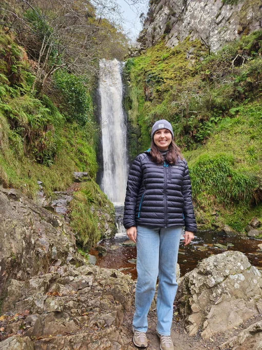 A person stands in front of a waterfall surrounded by lush greenery and rocky terrain. The individual is wearing a black puffer jacket and light blue jeans.