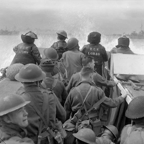 Black-and-white photo of Canadian soldiers packed into a landing craft on D-Day. Several soldiers wear helmets and heavy gear, with some facing forward toward rough water and a distant naval vessel. The backs of soldiers near the bow show markings on their jackets. Waves rise along the side of the craft as it moves through the water.
