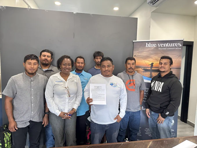 Belize Country Director Breanna Mossiah-Conorquie stands with a group of members from the San Estevan Fishers\u2019 Association in an office setting; one member holds up a document, representing their collaborative partnership with Blue Ventures.