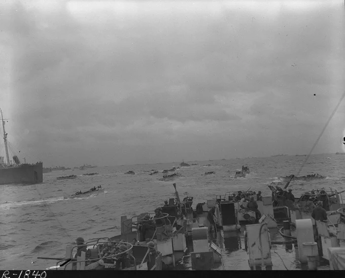 Numerous small boats filled with soldiers approach a large ship on a cloudy day during a military amphibious operation.
