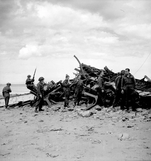 Soldiers in World War II gear stand and move around a large pile of twisted metal debris on a sandy beach.