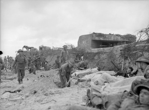 Soldiers in uniform on sandy ground beside a tall concrete wall topped with barbed wire during wartime.